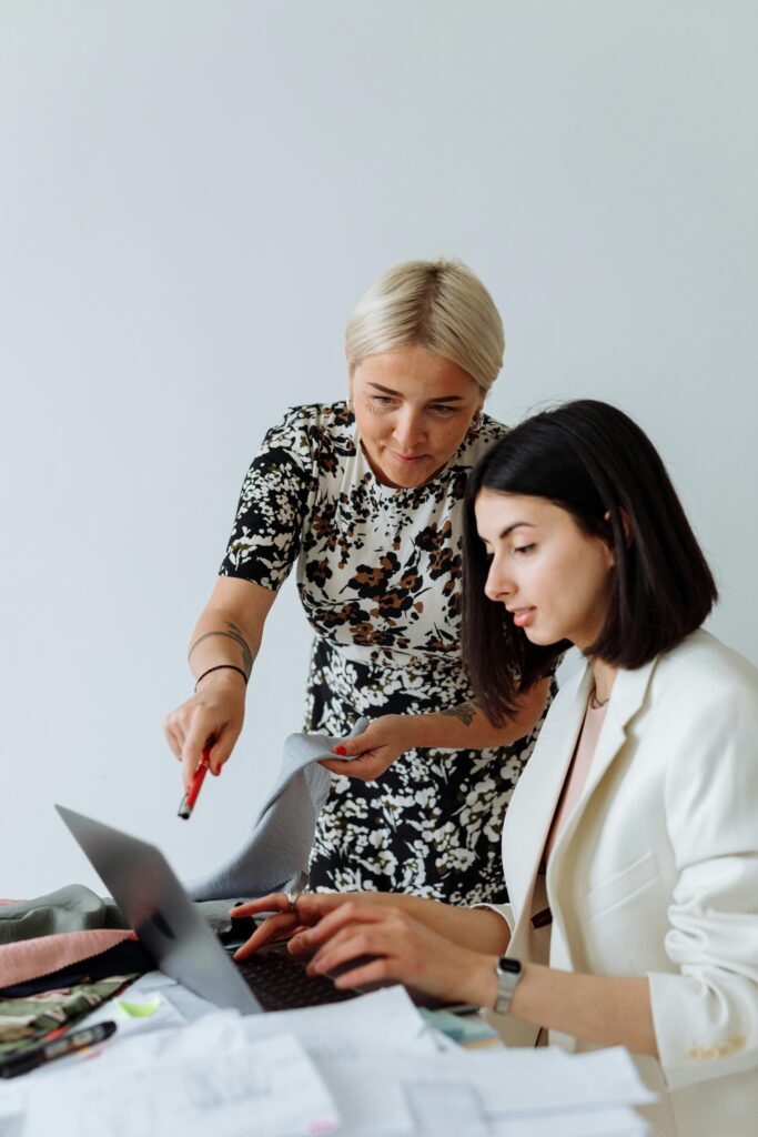 Two women discussing a project at a desk with a laptop in a modern office setting.